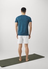 Hombre de pie descalzo sobre una esterilla de yoga oscura, de espaldas, vistiendo una camiseta azul y pantalones cortos blancos, contra un fondo blanco liso.