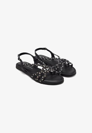 Sandalias negras con tiras cruzadas que presentan detalles metálicos, hebilla ajustable en el tobillo, suela plana y una plantilla de textura suave.