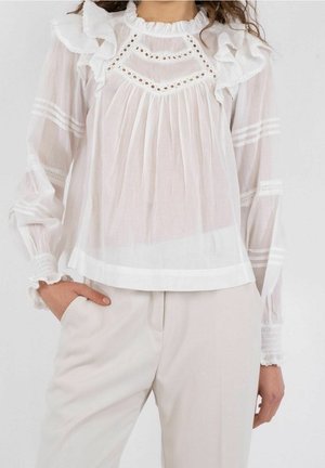 Femme portant un chemisier blanc transparent à volants et détails ajourés, manches longues, associé à un pantalon beige clair, main dans la poche.