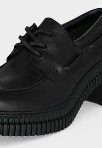 Chaussure montante en cuir noir avec semelle épaisse en caoutchouc crantée et coutures moc toe, montrée en gros plan sur fond blanc.