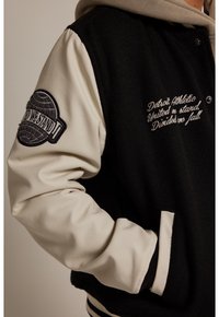 Giacca varsity nera e crema con corpo in lana, maniche in pelle, testo ricamato e toppa sulla manica. Vita a righe orizzontali.