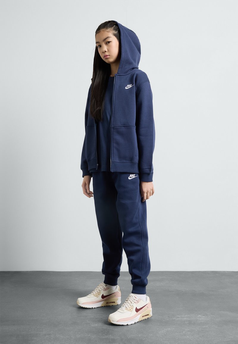 Marineblaue Zip-Hoodie mit einer Vordertasche, kombiniert mit passenden Jogginghosen. Die Schuhe haben eine Mischung aus hellen und burgunderfarbenen Akzenten.