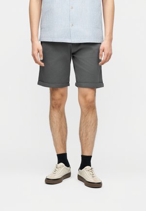 Mann trägt dunkelgraue hochgekrempelte Shorts, schwarze Socken, beige Turnschuhe und ein hellblaues kurzärmeliges Hemd, steht vor weißem Hintergrund.