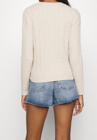 Maglione lavorato a maglia beige con motivo a treccia, maniche lunghe e scollatura rotonda, abbinato a pantaloni corti in denim blu con orlo sfrangiato.