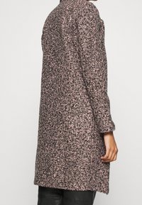 Persona con un abrigo largo de lana rosa y negro con textura y mangas largas, de pie contra un fondo claro y sencillo.