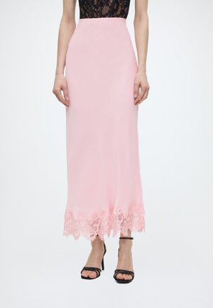 Γυναίκα που φοράει φούστα ροζ μήκους μέχρι το πάτωμα με δαντελένιο τελείωμα και μαύρα ψηλοτάκουνα πέδιλα με λουράκια, στέκεται μπροστά σε λευκό φόντο.