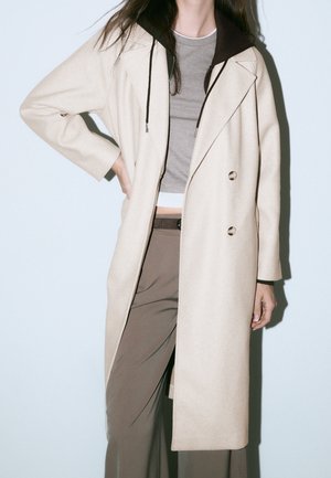 Persona con un abrigo largo beige sobre un top gris corto y pantalones marrones de cintura alta, de pie con una mano en la cadera contra un fondo claro.