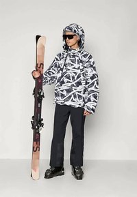 Εξοπλισμός σκι: ένα άτομο που φοράει ένα αδιάβροχο μπουφάν σε μαύρο και άσπρο σχέδιο, μαύρο παντελόνι, κρατάει σκι με βάσεις και μαύρες μπότες σκι.