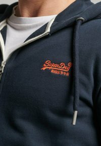 Felpa con zip blu navy e dettagli bianchi, caratterizzata da un logo ricamato arancione sul petto e una zip argentata.