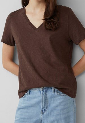 Kvinde iført en brun V-udskæring t-shirt med korte ærmer stoppet ned i lyseblå høj talje denim jeans, stående med hænderne bag ryggen.
