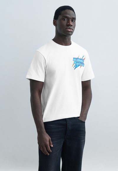 Άνδρας που φοράει λευκό μπλουζάκι με πολύχρωμο γραφικό λογότυπο στο στήθος, συνδυασμένο με σκούρα τζιν, στέκεται μπροστά σε ένα απλό γκρι φόντο.