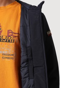 Giacca puffer blu navy con tessuto esterno liscio, dotata di zip completa, cappuccio e logo ricamato. Sotto, una t-shirt grafica arancione.