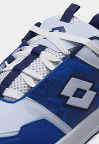 Λευκό και μπλε αθλητικό παπούτσι με λευκά κορδόνια, γλωττίδα από δίχτυ, σκούρες μπλε λεπτομέρειες και γεωμετρικό λογότυπο στην πλευρική επιφάνεια.
