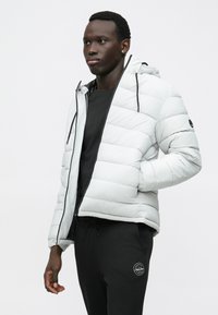 Ανοιχτό γκρι φουσκωτό μπουφάν με κουκούλα, χαρακτηρισμένο από οριζόντιες ραφές και μαύρες λεπτομέρειες στο φερμουάρ, συνδυασμένο με μαύρες φόρμες.