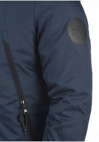 Temno modra jakna z teksturirano tkanino, ki ima stransko zadrgo in krožno črno oznako na levem rokavu.