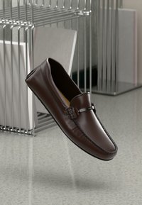 Mocassins en cuir marron avec une finition lisse, dotés d'une sangle avant avec un accent métallique et d'une couture détaillée le long des bords.