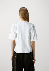 Weißes Baumwoll-T-Shirt mit kurzen Puffärmeln und Verzierungen an den Schultern. Lässiger Schnitt mit rundem Halsausschnitt, glatte Textur.