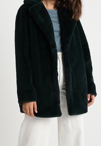 Tummanvihreä tekoturkistakki huivikurilla. Siinä on pitkät hihat, avoin etuosa ja sileä pinta, käytetty vaaleansinisen topin päällä.