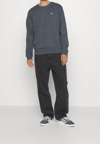 Sudadera de cuello redondo azul oscuro con puños acanalados, combinada con jeans de pierna ancha negros y zapatillas oscuras con rayas blancas.
