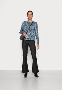 Blusa azul de mangas compridas com padrão abstrato preto, combinada com calças pretas de cintura alta e botas de cano curto pretas com salto. Mala preta com alça para o ombro.