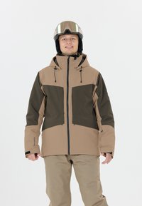 Giacca da sci beige e verde scuro con chiusura a zip, cappuccio regolabile, polsini elasticizzati e pantaloni beige abbinati. Il modello indossa occhiali da sci.