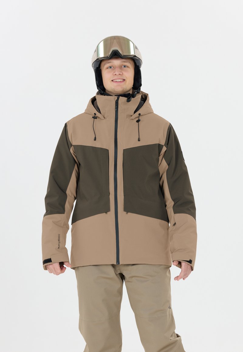 Giacca da sci beige e verde scuro con chiusura a zip, cappuccio regolabile, polsini elasticizzati e pantaloni beige abbinati. Il modello indossa occhiali da sci.