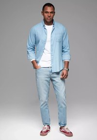Lichtblauwe overhemdo over een wit t-shirt, gecombineerd met lichtblauwe spijkerbroek en rode sneakers; textuur van het shirt, opgerolde mouwen en horloge.