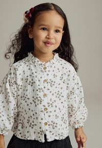 Vit skjorta med kort ärm och liten blommönster, volangkrage och puffärmar. Har träknappar och mjukt, strukturerat tyg.