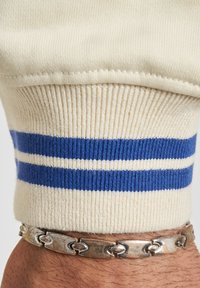 Kremfarget strikket ermekant med to blå striper og en hvit stripe. Et sølvfarget kjedearmbånd viser et polert, sammenkoblet design mot huden.