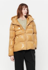 Giovane donna che indossa un lucido giubbotto imbottito color sabbia sopra una camicia verde chiaro con bottoni e pantaloni neri, in piedi su uno sfondo bianco semplice.