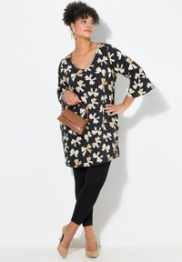 Tunic nera a fiori con motivi beige, maniche a campana a tre quarti, abbinata a leggings neri e tacchi beige. Include una piccola borsa marrone.