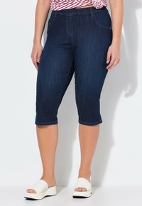 Pantalones capri de mezclilla oscura con un diseño ajustado, cintura de corte medio y dobladillo enrollado, combinados con sandalias de plataforma blancas.