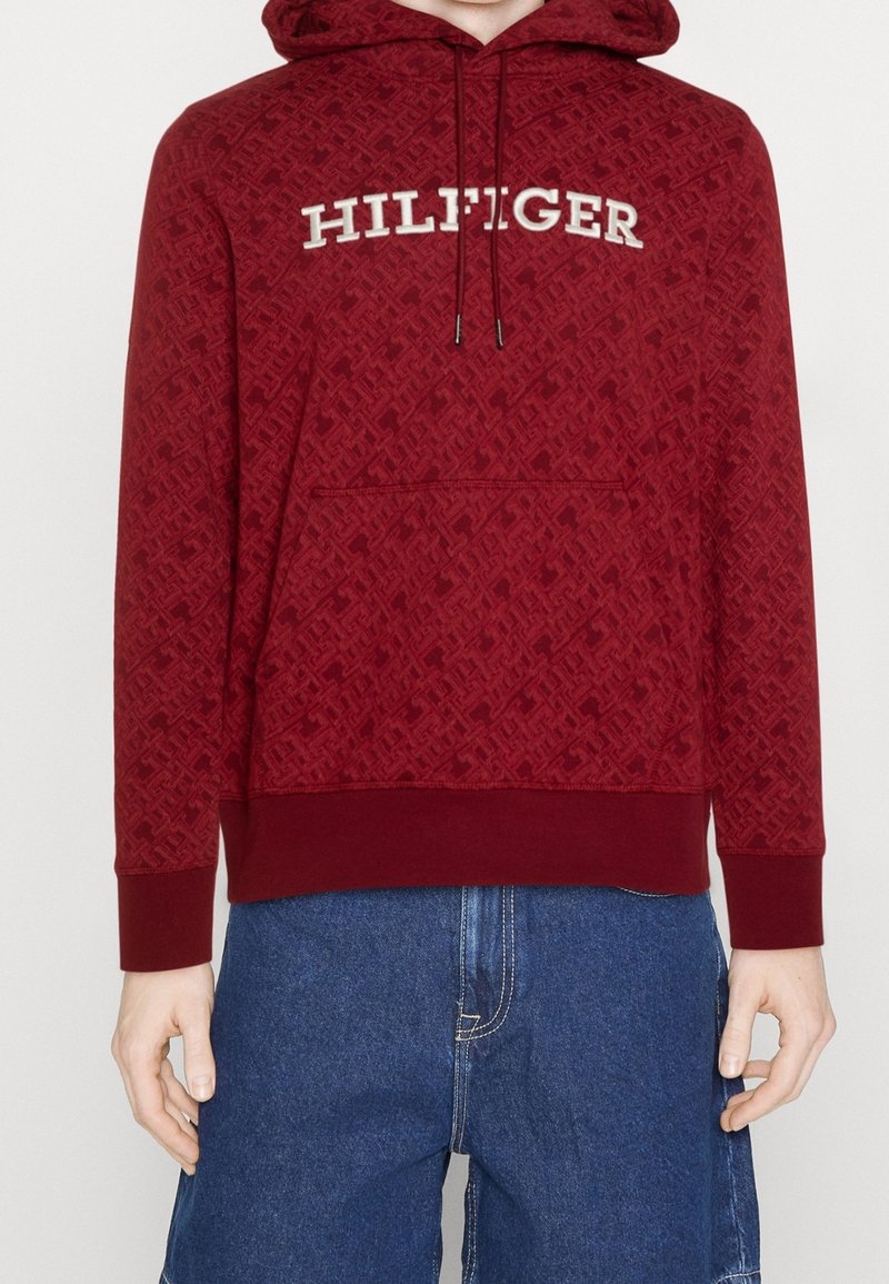 Vue de face d'une personne portant un sweat à capuche rouge texturé avec le texte "HILFIGER" et un jean en denim bleu, les mains détendues le long du corps.