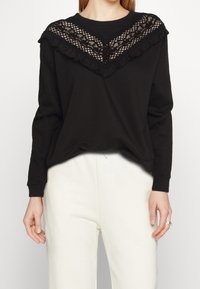 Sudadera negra con un diseño de encaje y volantes en el escote. Tejido suave, corte relajado, con mangas largas y un sutil patrón texturizado.
