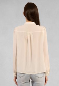 Camicia trasparente color crema con maniche lunghe, polsini a bottone e un design posteriore plissettato. La texture setosa esalta l'aspetto leggero e fluido.
