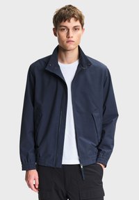 Jonge man met een marineblauwe jas met rits over een wit shirt en zwarte cargobroek, staand tegen een effen lichte achtergrond.