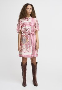 Rochie roz cu un model, cu mâneci scurte, guler și un cordon la talie. Asortată cu cizme din piele maro până la genunchi. Textura materialului este netedă.