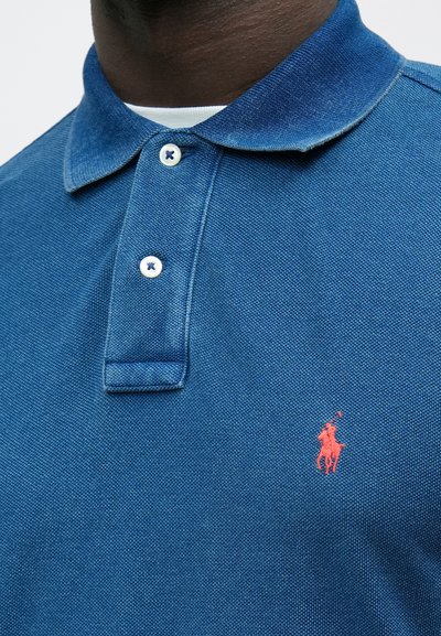 Modra polo majica iz teksturirane tkanine s kratkimi rokavi, s poklopcem na ovratniku z gumbi in majhnim rdečim logotipom na prsih.