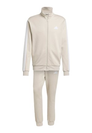 Bežs Adidas treniņtērps ar rāvējslēdzēju, kas ietver jaku un atbilstošas bikses. Uz rokām ir baltas svītras un uz abiem izstrādājumiem ir logo akcenti. Maiga tekstūra.