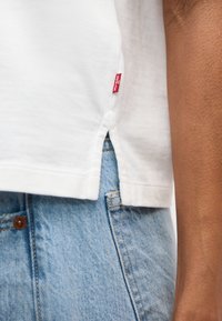 Λευκός ρεβέρ βαμβακερού πουκάμισου με κόκκινη ετικέτα Levi's πάνω από ανοιχτόχρωμο τζιν τζιν σε μέση προσώπου, με ορατό το χέρι στα δεξιά.