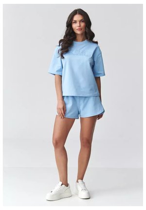 Kobieta z długimi ciemnymi włosami, ubrana w jasnoniebieski oversize komplet t-shirt i szortów, stoi z jedną ręką luźno opuszczoną, białe sneakersy na platformie.