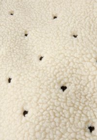 Texture de tissu en polaire de couleur crème avec de petits points marron foncé uniformément espacés. Surface douce, moelleuse et légèrement texturée.