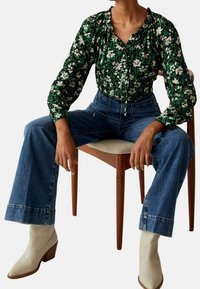 Person, der sidder på en træstol iført en grøn blomsterbluse, højtaljede, bredbenede blå jeans og hvide ankelstøvler.