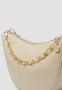 Cremefarbene Lederhandtasche mit geschwungener Form, goldfarbener Kettenriemen und einem Reißverschluss oben. Enthält einen dekorativen Logo-Anhänger.