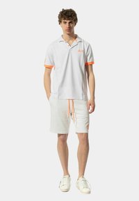Polo bianco con accenti arancioni e logo, abbinato a pantaloncini bianchi con coulisse arancione, indossato con sneakers bianche.
