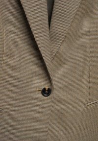 Hnědý saka s kostkovaným vzorem houndstooth, textura látky, s černým knoflíkem a klopami, obsahující jemné švy.