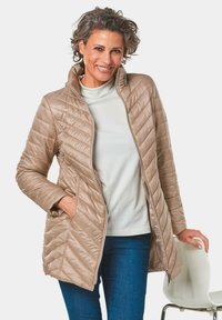 Beige pufferjack met diagonaal doorgestikte patroon, ritssluiting en hoge kraag, gedragen over een lichtgekleurde top en een spijkerbroek.