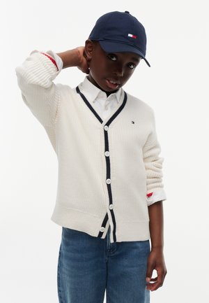 Garçon portant une casquette bleu marine, un cardigan blanc boutonné avec bordure bleu marine, une chemise blanche et un jean bleu, touchant l'arrière de son cou.