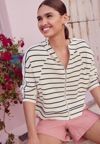 Witte zip-up hoodie met marineblauwe strepen, gestructureerde stof en raglanmouwen. Gecombineerd met roze shorts. Achtergrond met roze muren en bloemen.