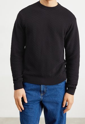 Person iført en sort, struktureret langærmet sweater og blå jeans, stående mod en ensartet lys baggrund.